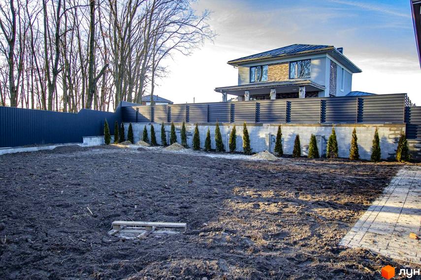 Котеджне містечко КМ NOVYI SVIT: двоповерховий будинок з цегляними вставками та сірим фасадом, огороджений парканом, з висадженими туями та підготовленою ділянкою для ландшафтного дизайну.