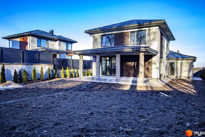 Двоповерховий будинок КМ NOVYI SVIT зі світлим фасадом та темним дахом. Прибудинкова ділянка з підготовленим ґрунтом та рядами туй біля паркану.