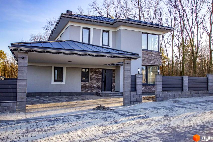 Двоповерховий будинок Котеджного містечка NOVYI SVIT з сірим фасадом, цегляними вставками та навісом для авто. Прибудинкова територія вимощена бруківкою, огороджена парканом з блоків та панелей.