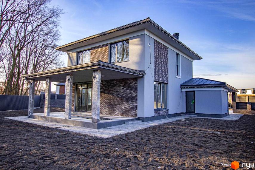 Двоповерховий будинок Котеджного містечка NOVYI SVIT з сірим фасадом та цегляними вставками. Перед будинком ґрунтова ділянка та вимощена доріжка.