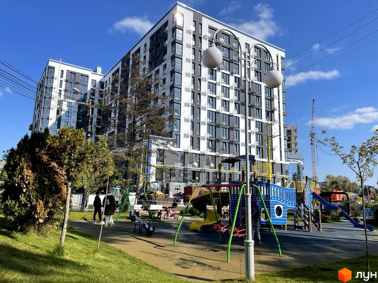 Новобудова ЖК Рів'єра з біло-сірим фасадом та дитячим майданчиком на прибудинковій території. Будівельний проєкт з сучасною архітектурою та зоною відпочинку.