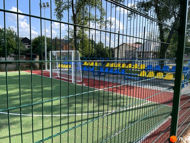 Спортивний майданчик з футбольними воротами та трибунами для глядачів у житловому комплексі Рів'єра. Зелене покриття поля та синьо-жовті сидіння створюють яскраву атмосферу.