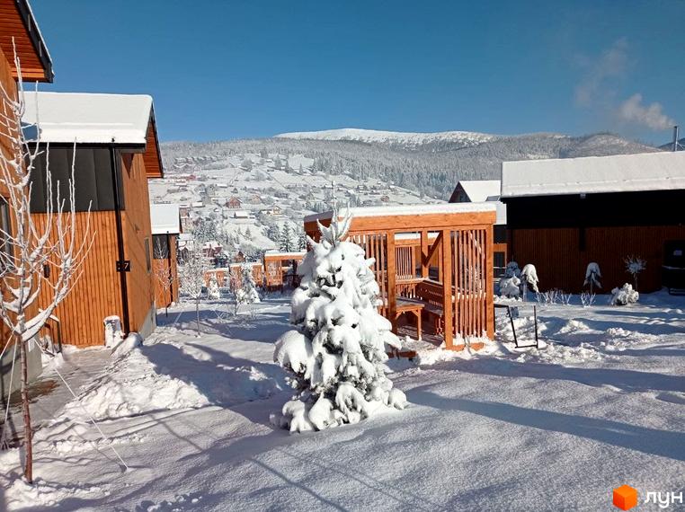 Зимовий пейзаж Котеджного містечка Phoenix Relax Park: дерев'яні котеджі та засніжена територія з ялинками. На задньому плані видніються гори.