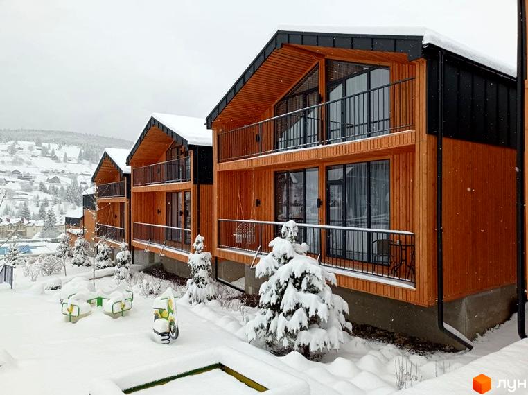 Засніжені котеджі Котеджного містечка Phoenix Relax Park з дерев'яними фасадами та балконами. Територія покрита снігом, є ялинки та дитячі гойдалки.