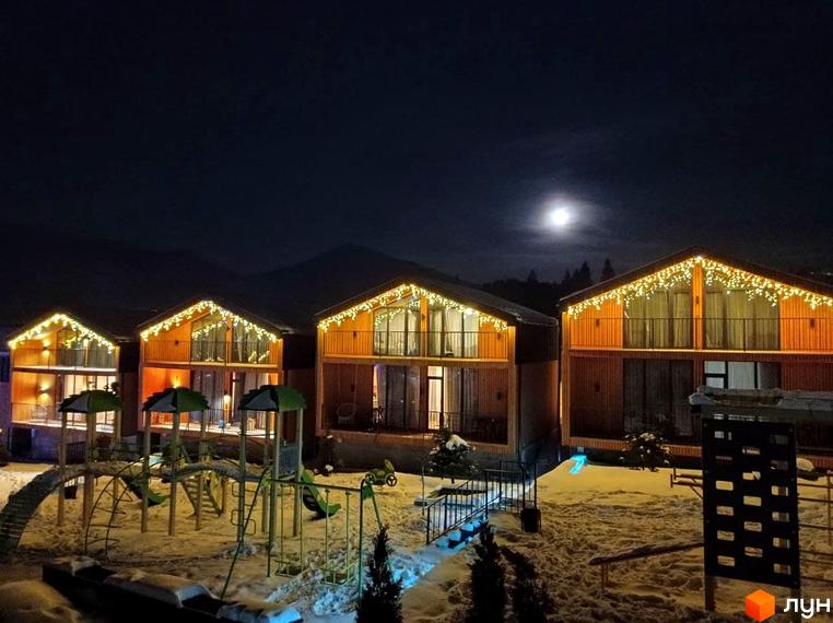 Вигляд на Котеджне містечко Phoenix Relax Park вночі. Будинки з дерев'яним оздобленням та ілюмінацією, дитячий майданчик взимку.