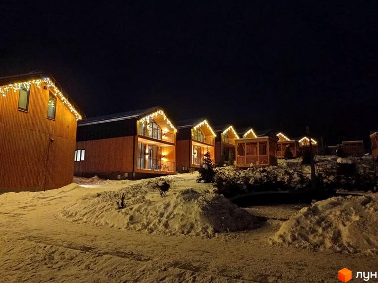 Зображення Котеджного містечка Phoenix Relax Park вночі. Дерев'яні будинки з гірляндами та засніжена територія.