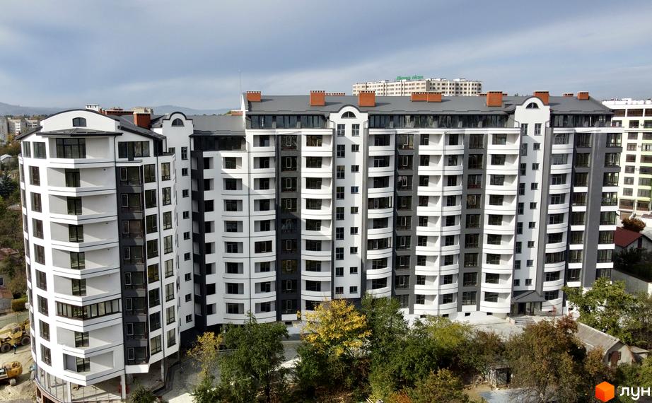 Новобудова Трускавецька Фортеця: багатоповерховий будинок з біло-сірим фасадом, вигнутими секціями та численними вікнами. Навколо видно дерева.