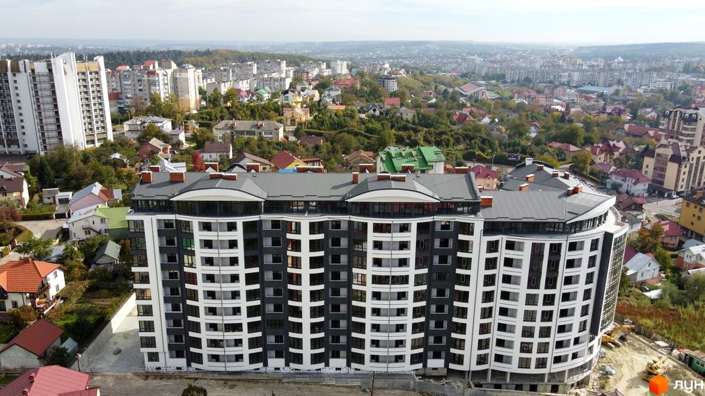 Житловий комплекс Трускавецька Фортеця: багатоповерхова будівля з вигнутим фасадом у білих та темно-сірих тонах. Навколо видно приватні будинки та міський пейзаж.