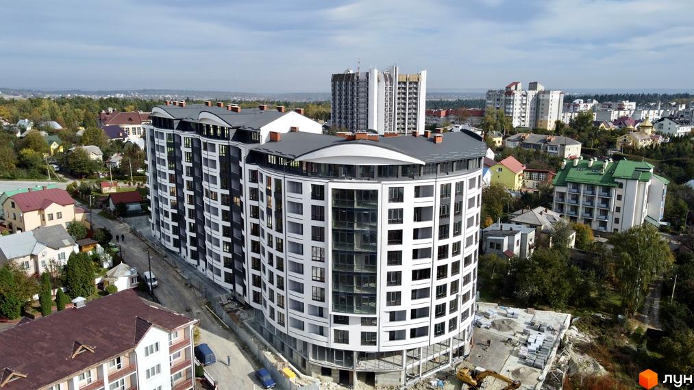 Новобудова Трускавецька Фортеця: багатоповерховий житловий будинок з біло-темним фасадом та вигнутою формою, поруч з приватною забудовою.