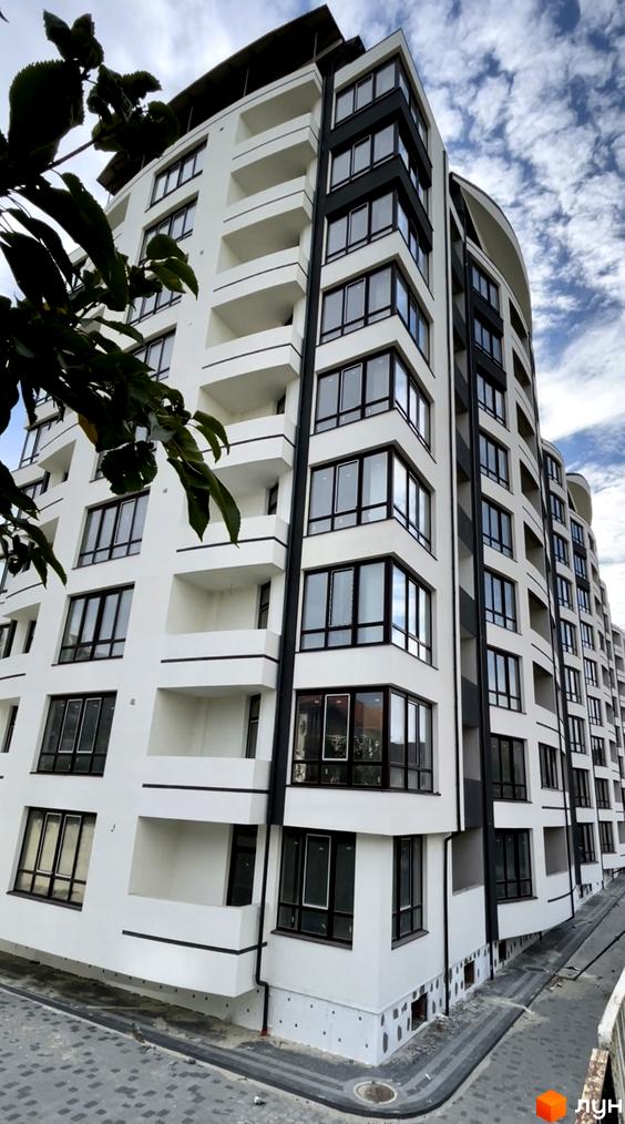 Житловий комплекс Трускавецька Фортеця: багатоповерховий будинок з білим фасадом, темними вікнами та балконами. Прибудинкова територія вимощена бруківкою.