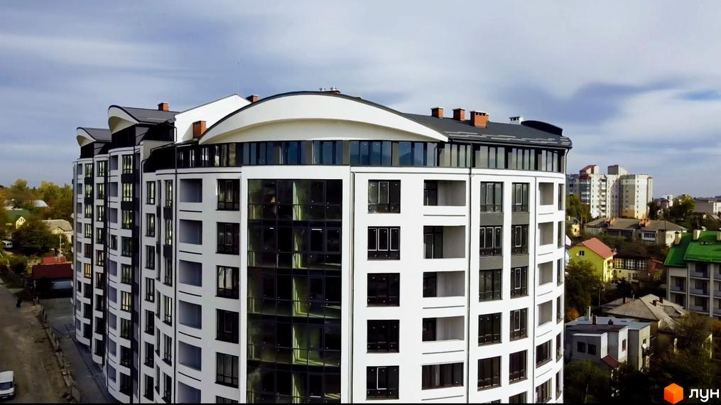 Новобудова Трускавецька Фортеця з білим фасадом, темними вікнами та округлими елементами даху. Навколо видно інші будівлі та дерева.