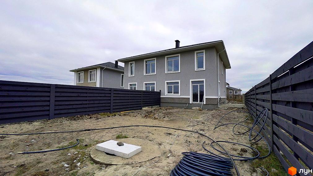 Зовнішній вигляд двоповерхового будинку сірого кольору з білими вікнами та темною огорожею. На прибудинковій території видно будівельні матеріали та труби.