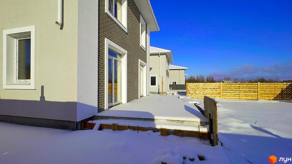 Зовнішній вигляд будинку з сірою цегляною обробкою та білими елементами фасаду. Навколо будинку засніжена територія та дерев'яний паркан.