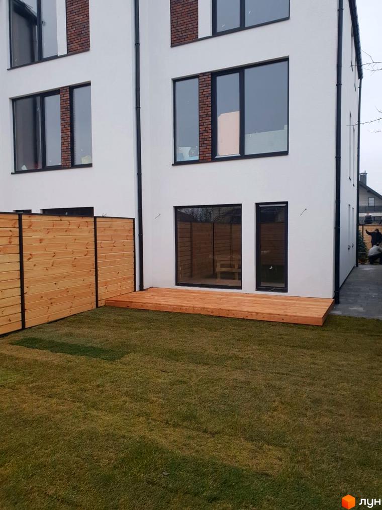 Таунхауси DOMA NOVA з великими вікнами та дерев'яною терасою. Навколо будинку зелений газон та дерев'яний паркан.