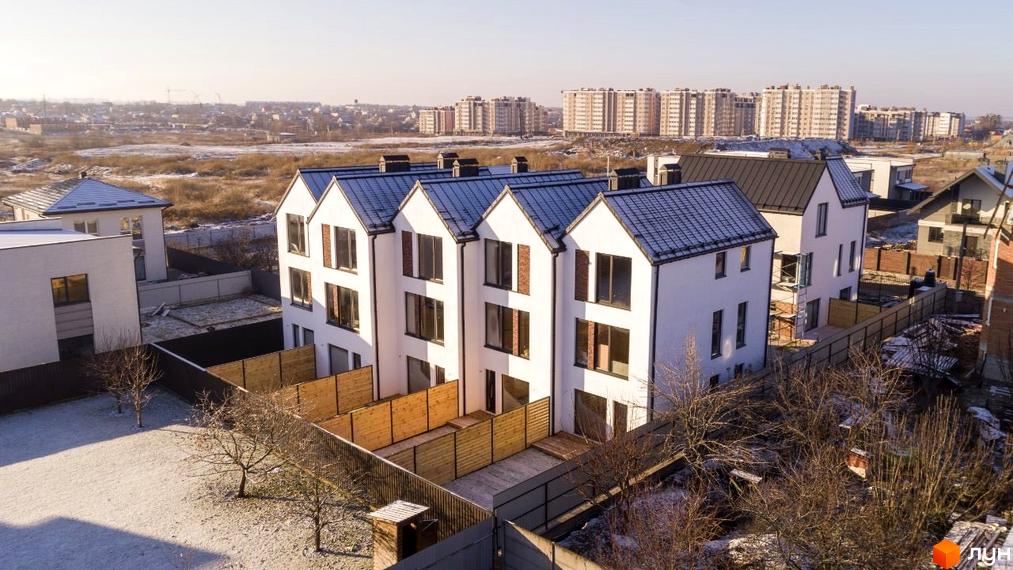 Таунхауси DOMA NOVA з білими фасадами та темними дахами на тлі міської забудови. Прибудинкова територія з парканом та деревами.
