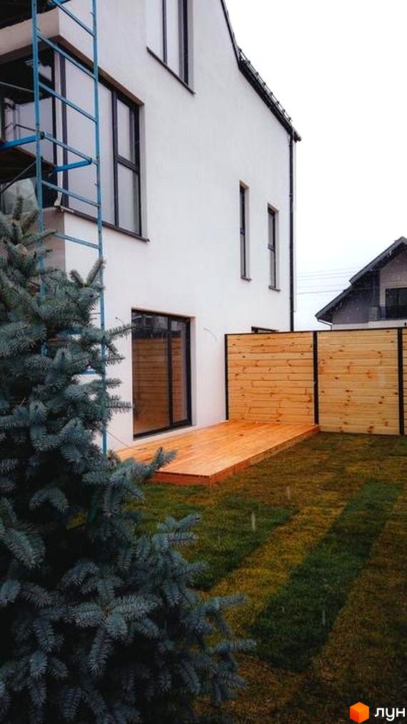 Таунхауси DOMA NOVA: білий двоповерховий будинок з дерев'яною терасою та огорожею. Навколо будинку зелений газон та хвойне дерево.