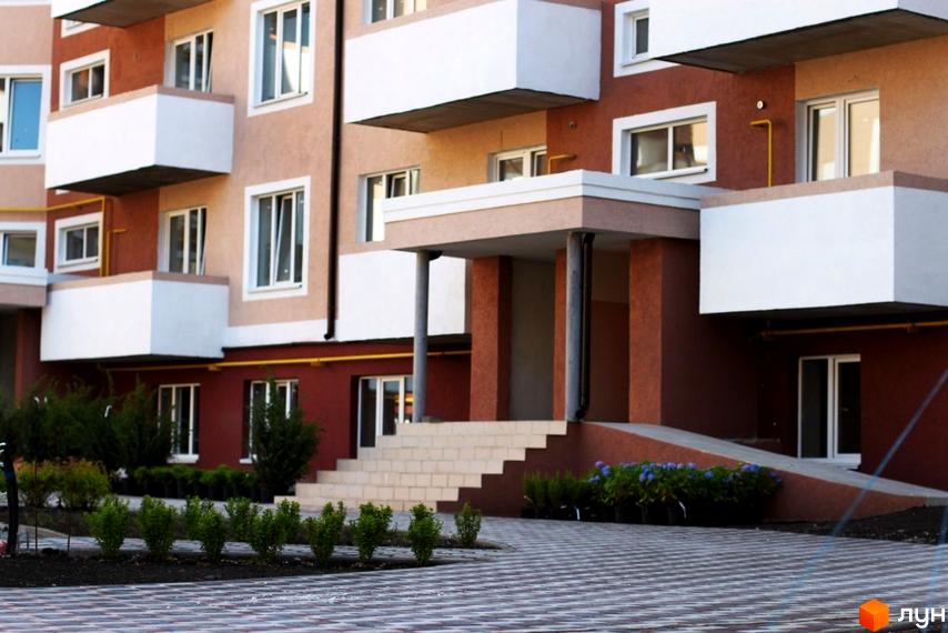 Житловий комплекс Новатор з фасадом у світлих тонах та балконами. Прибудинкова територія з озелененням та плитковою доріжкою.