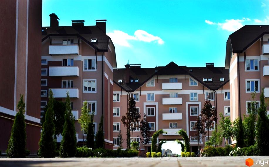 Зовнішній вигляд житлового комплексу ЖК Новатор з фасадами світло-бежевого кольору та темними дахами. Прибудинкова територія з зеленими насадженнями та арками.