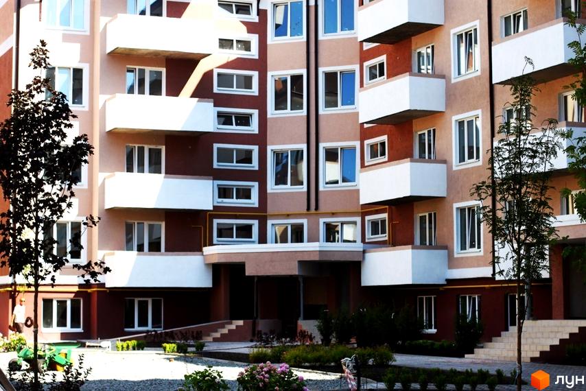 Житловий комплекс Новатор – багатоповерховий будинок з білими балконами та коричневими фасадами. На прибудинковій території розташовані дерева та зелені насадження.