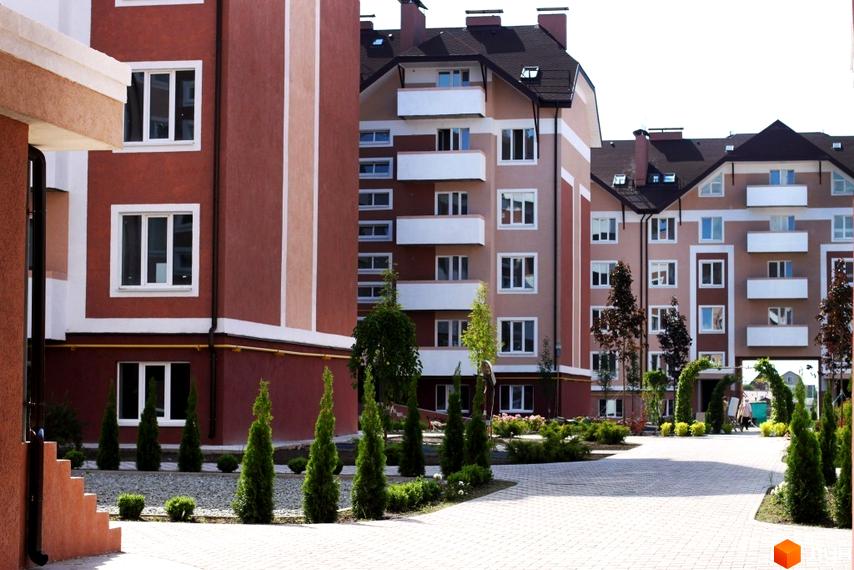 Зовнішній вигляд ЖК Новатор з фасадами в бордових та бежевих тонах, балконами та вікнами. Прибудинкова територія з туями, кущами та бруківкою.