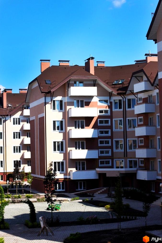 Житловий комплекс Новатор з коричневими дахами та світлими фасадами. На прибудинковій території розташовані дерева та доріжки.