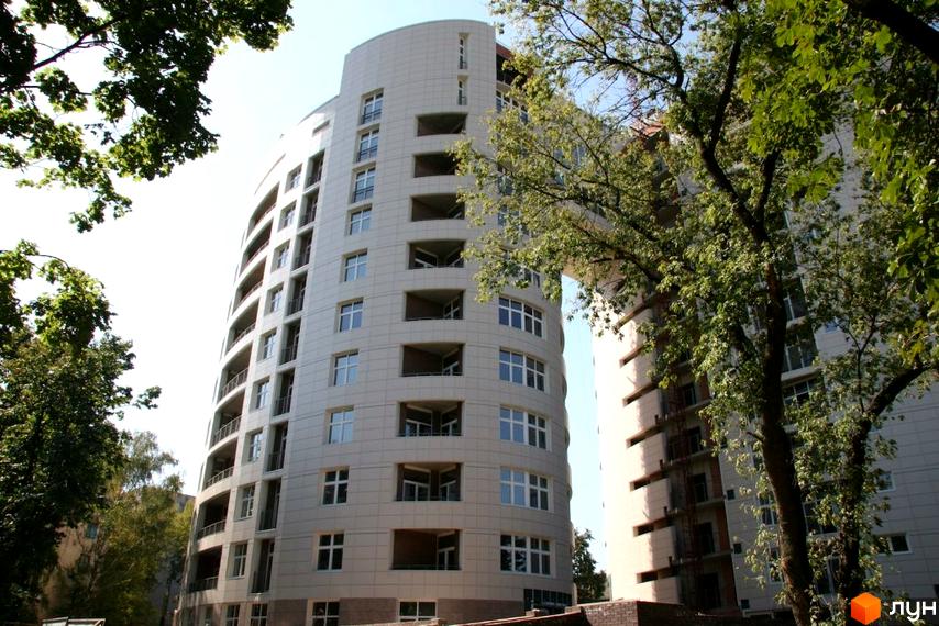 Житловий комплекс Схід-Захід - це двосекційна новобудова з білою фасадною обробкою та панорамними вікнами, оточена зеленню дерев.