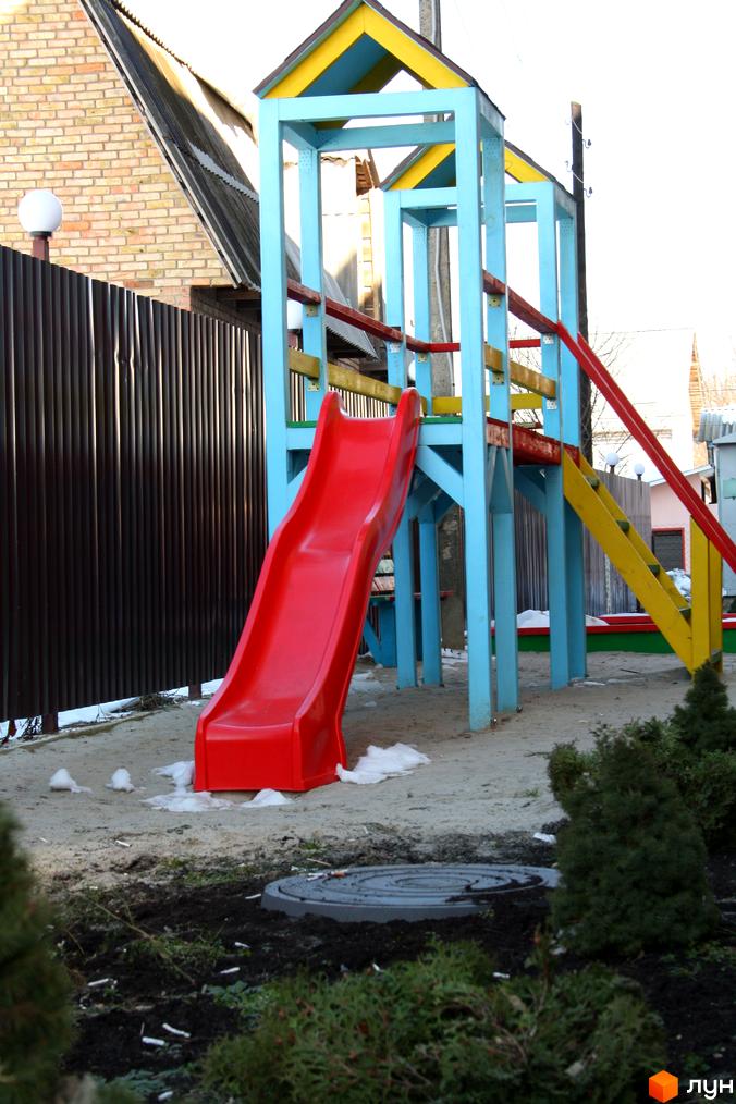 Дитячий майданчик з червоною гіркою та кольоровою конструкцією, оточений зеленими насадженнями на прибудинковій території.