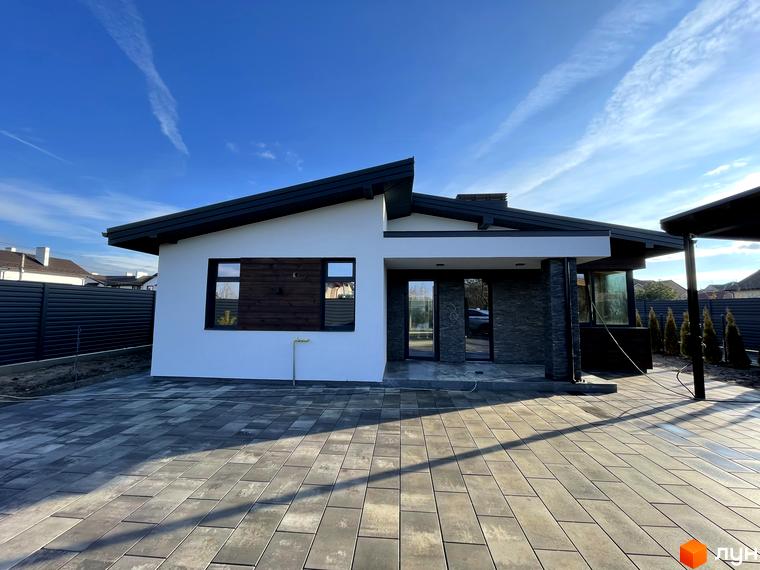 Новобудова: одноповерховий будинок з білими стінами, темним дахом та кам'яною обробкою, перед яким вимощена сіра бруківка.