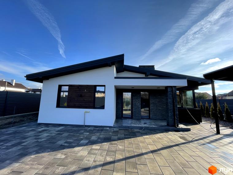 Сучасний одноповерховий будинок з білою штукатуркою, темним дахом та кам'яним оздобленням фасаду. Прибудинкова територія вимощена сірою плиткою.