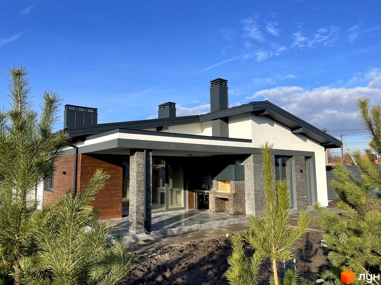 Одноповерховий будинок з темно-сірим дахом, білими та дерев'яними фасадами, критою терасою і зоною барбекю. Навколо молоді сосни та підготовлена ділянка на вул. Мазепи.