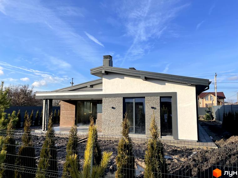 Одноповерховий будинок зі світлим фасадом, темним дахом та кам'яним оздобленням. Прибудинкова територія з молодими хвойними деревами.