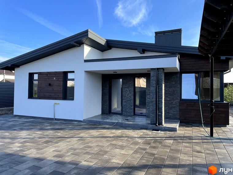 Одноповерховий будинок з комбінованим фасадом: білі стіни, дерев'яні панелі та кам'яне облицювання. Подвір'я вимощене бруківкою.