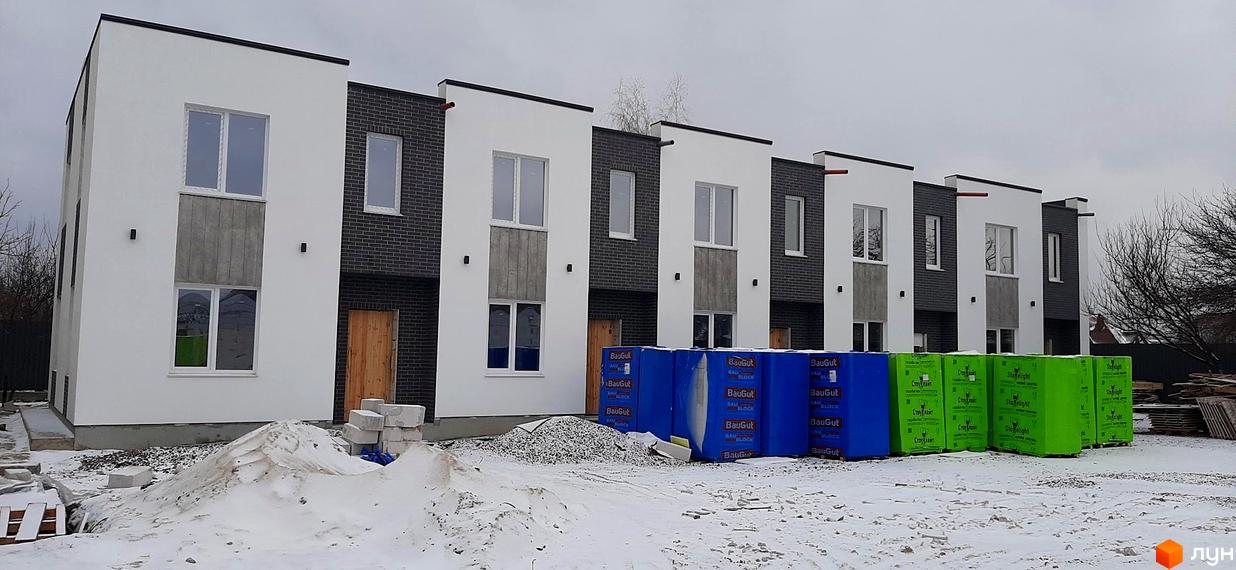 Зовнішній вигляд будинків житлового комплексу КМ Holland Park з білим та сірим фасадом, вікнами та дерев'яними дверима. На передньому плані видно будівельні матеріали та сніг.