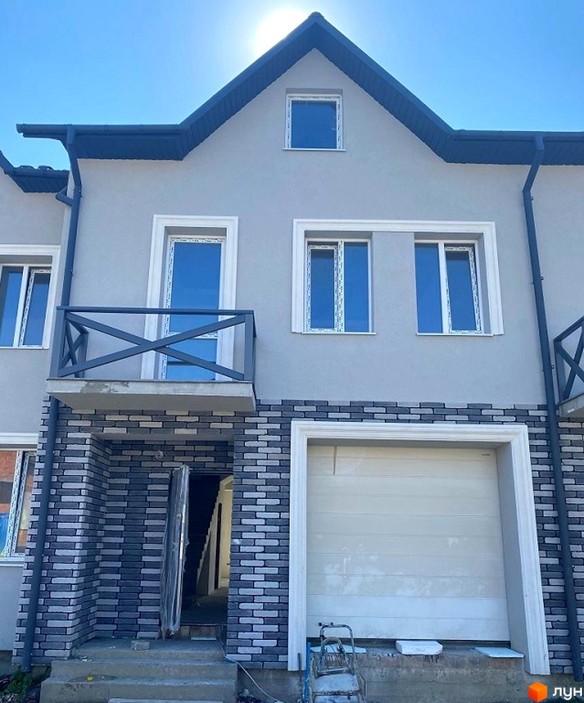 Зовнішній вигляд таунхаусів Hodillywood 2.0 з сірим фасадом, цегляним цоколем та гаражними воротами. Навколо будинку видно елементи прибудинкової території.