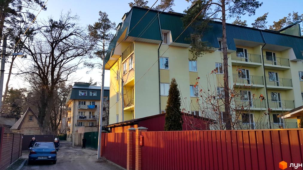 Житловий комплекс Теплий дім має світло-жовтий фасад, зелений дах та балкони. Навколо розташовані високі дерева, червоний паркан і ґрунтова дорога.