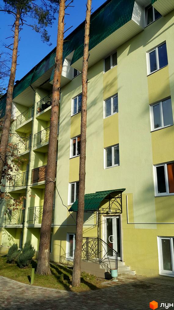 Зовнішній вигляд житлового комплексу Теплий дім з жовтою фасадною обробкою та зеленим дахом. Приблизно біля будинку ростуть сосни та зелена трава.