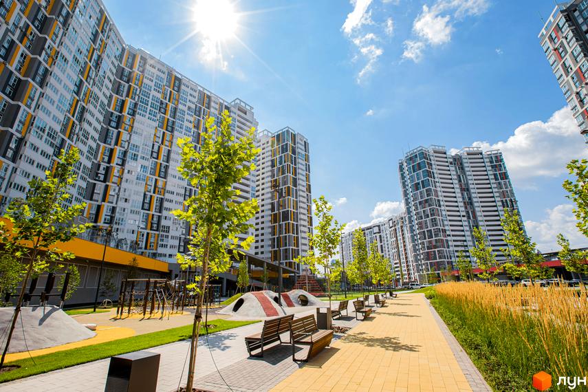 Житловий комплекс Русанівська Гавань: високі будинки з білими, сірими та жовтими фасадами, доріжки, лавки та дитячий майданчик на прибудинковій території.