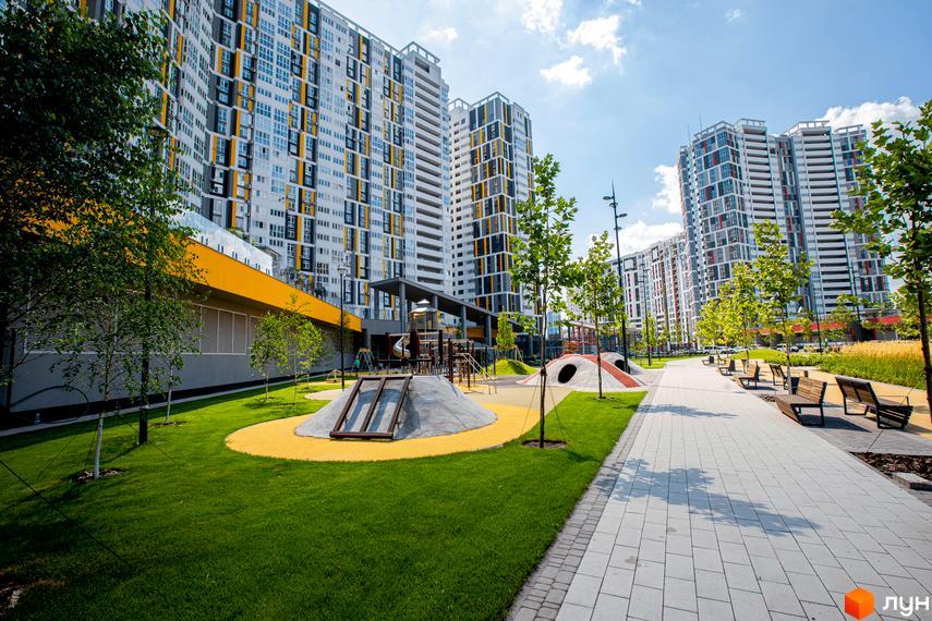 Високі будинки ЖК Русанівська Гавань з фасадами у світлих тонах. На території комплексу дитячий майданчик, зелені газони та доріжки для прогулянок.