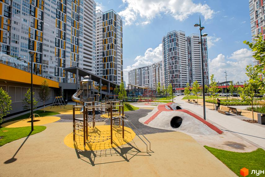 Багатоповерхові житлові будинки з білими, сірими та жовтими фасадами та облаштований дитячий майданчик з гіркою і лазалками на території житлового комплексу Русанівська Гавань.