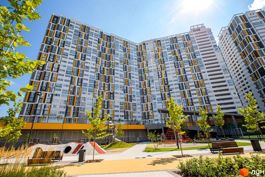 Висотна будівля ЖК Русанівська Гавань з фасадом білого, жовтого та сірого кольорів. На території комплексу дитячий майданчик та зелені насадження.