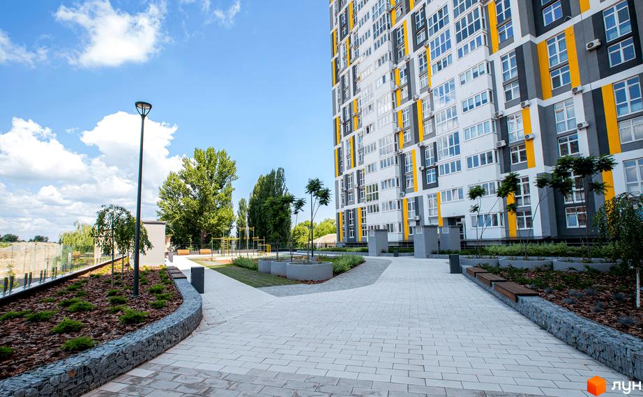 Висотна будівля житлового комплексу з білими, сірими та жовтими фасадами. На передньому плані доглянута прибудинкова територія з бруківкою, деревами та лавками.