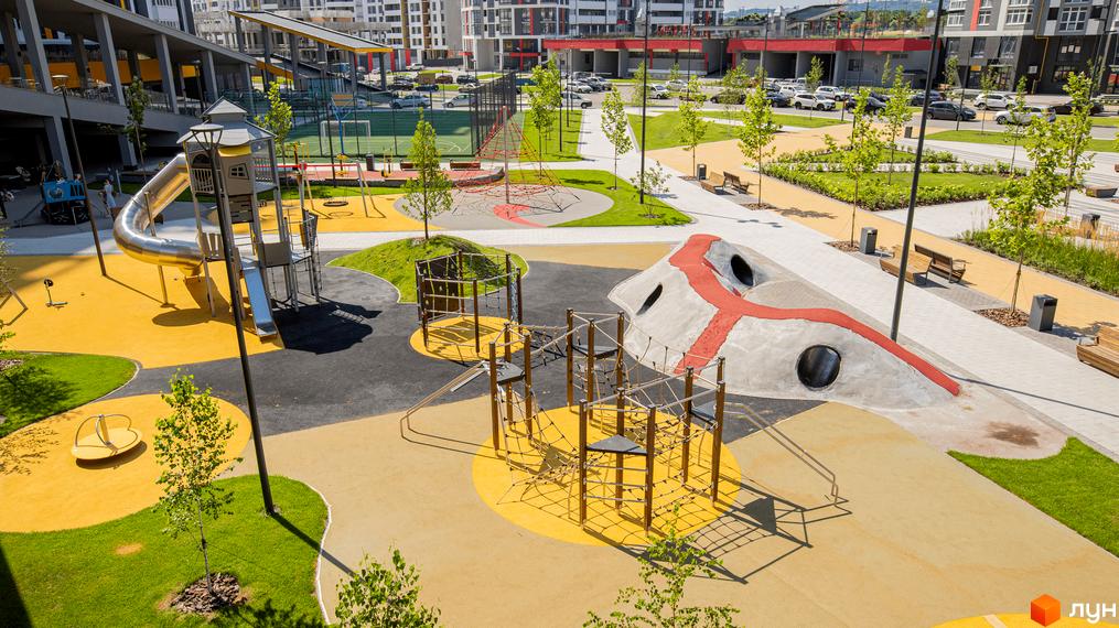 Житловий комплекс Русанівська Гавань з багатоповерховими будинками та просторим дитячим майданчиком з різноманітними ігровими елементами, зеленими газонами та доріжками.