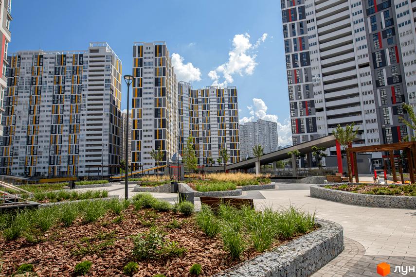 Високі житлові будинки з білими фасадами, сірими та жовтими вставками. На передньому плані - доглянута прибудинкова територія з газонами та декоративним камінням.