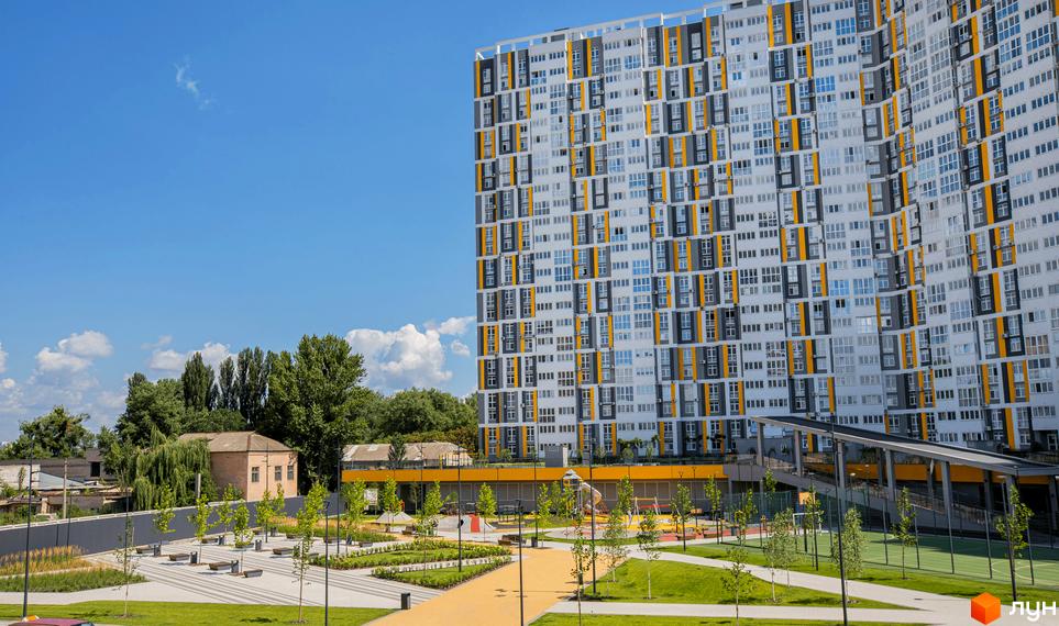 Багатоповерховий житловий будинок Русанівська Гавань з біло-сірим фасадом та жовтими акцентами. Прибудинкова територія має зелені насадження, доріжки та спортивний майданчик.