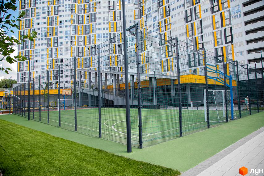 Багатоповерховий будинок ЖК Русанівська Гавань з фасадом у світлих тонах. На території розташований спортивний майданчик із зеленим покриттям та огорожею.