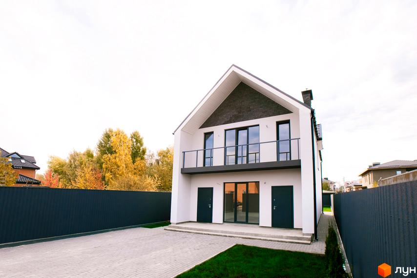 Білий двоповерховий будинок Котеджного містечка Щастя з темним скатним дахом та балконом. Перед будинком вимощена територія, зелений газон та темний паркан.