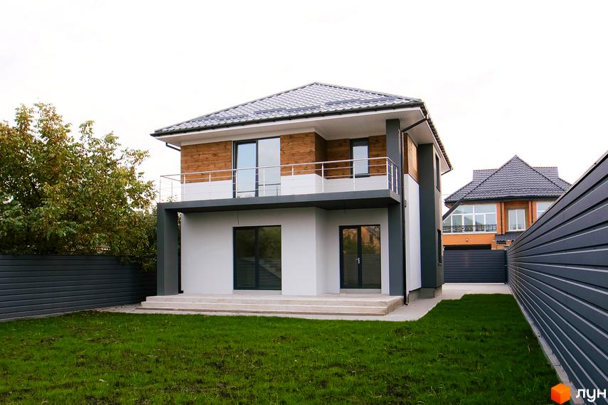 Двоповерховий будинок з дерев'яним оздобленням фасаду та балконом. На передньому плані газон та огорожа, на задньому - сусідній будинок.