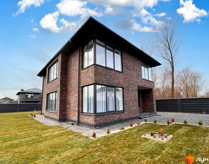 Двоповерховий будинок з цегляною кладкою та великими вікнами. На прибудинковій території газон, декоративні кущі та бруківка.
