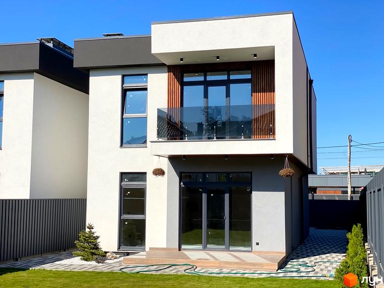 Двоповерховий будинок в Котеджному містечку Щастя з великими вікнами та балконом. На подвір'ї газон, доріжки з бруківки та невеликі дерева.