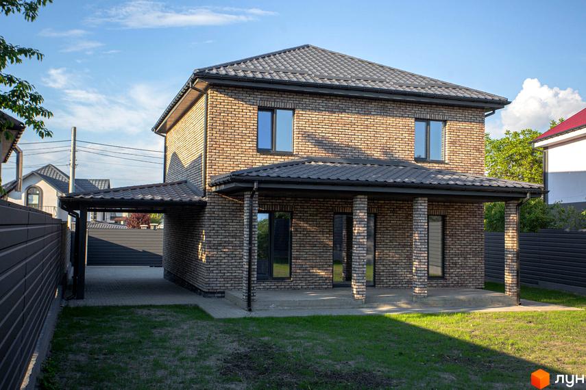 Двоповерховий цегляний будинок Котеджного містечка Щастя з темно-сірим дахом та навісом для авто. Перед будинком зелений газон.